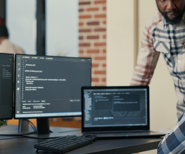 Focused software developer writing code on laptop looking at multiple screens with programming language is interrupted by colleague coder asking for advice. Programmers doing online cloud computing.